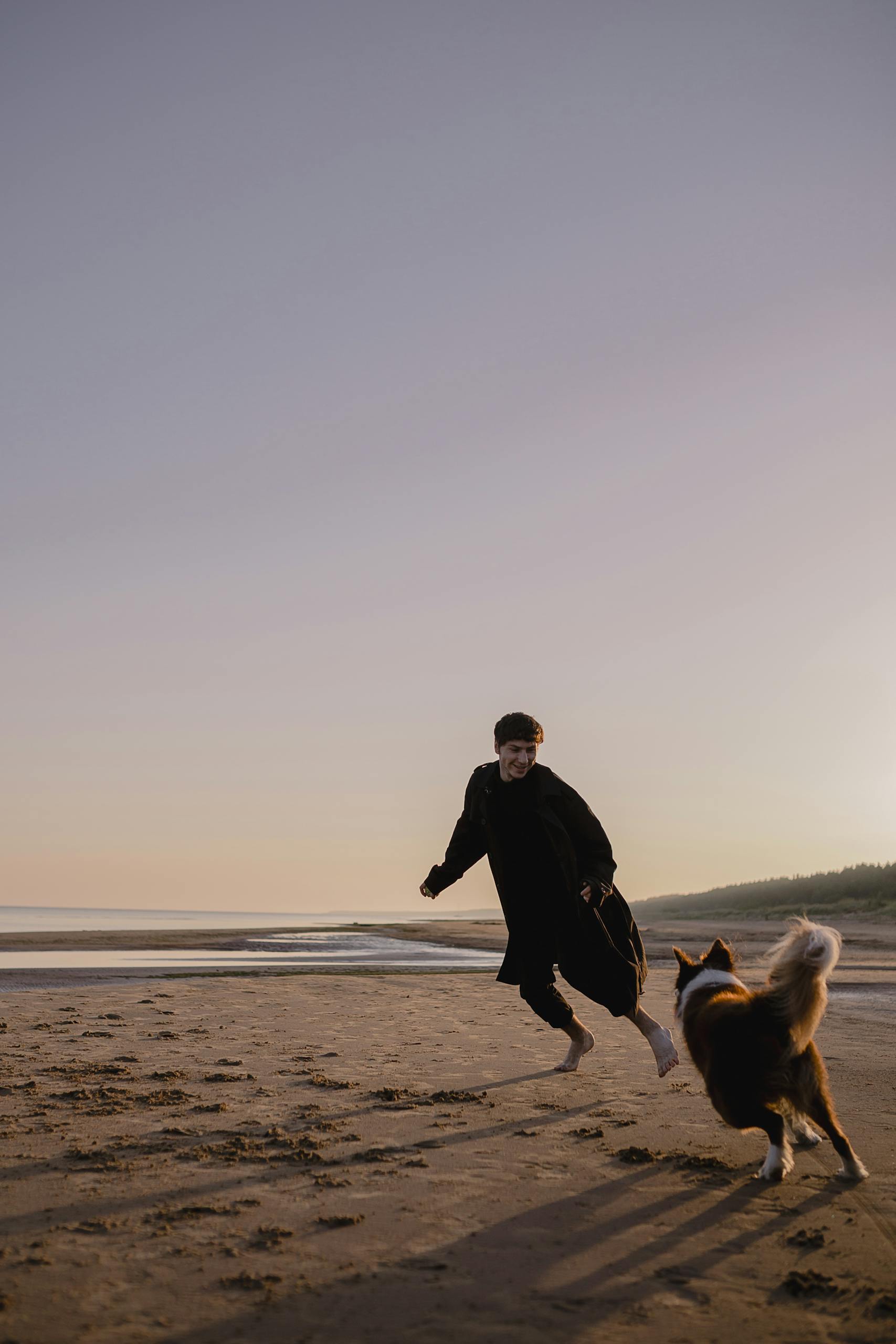 A man joyfully runs barefoot on the beach with his dog during a beautiful sunset.