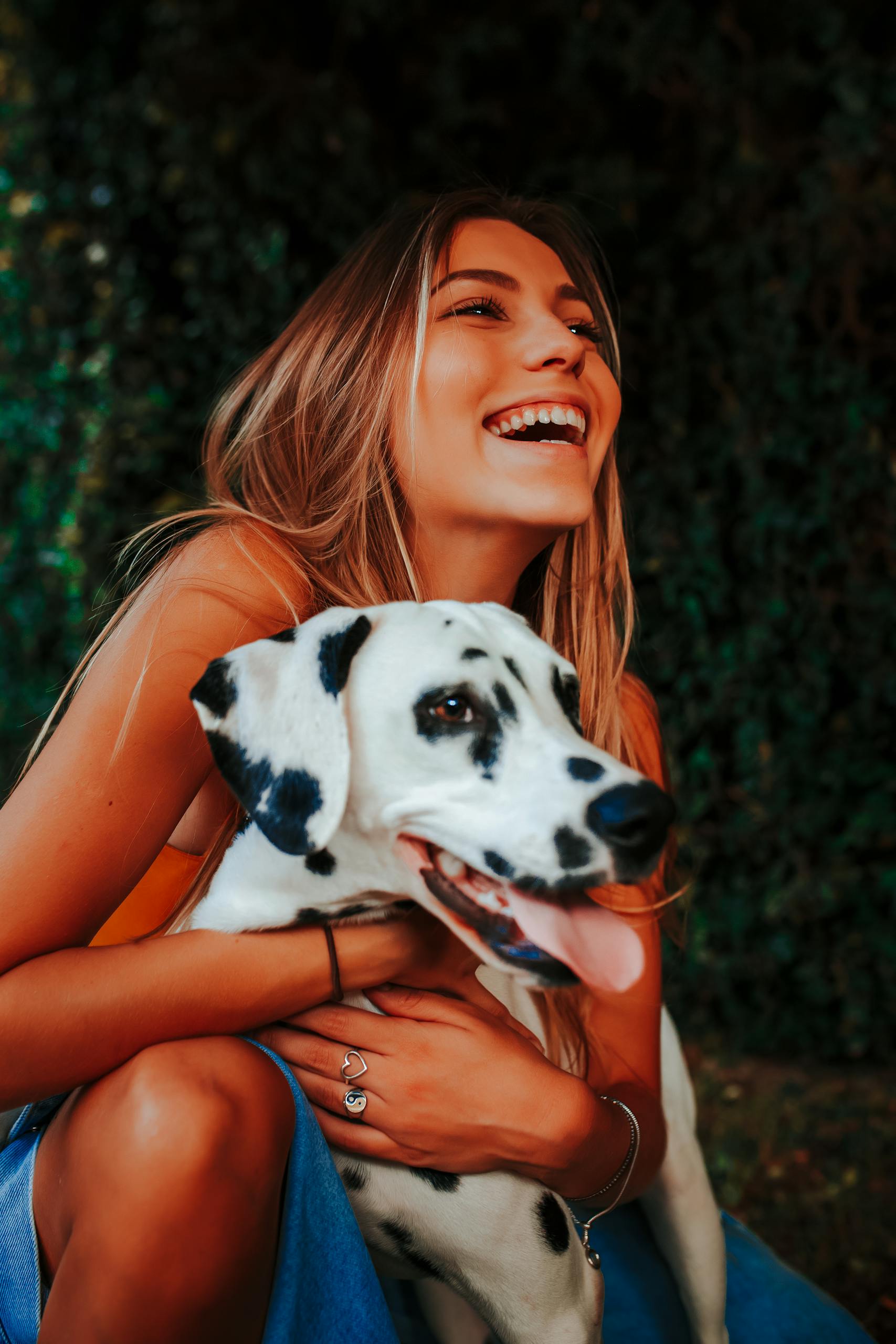 A cheerful woman with long blonde hair embracing her happy Dalmatian dog outdoors on a bright day.