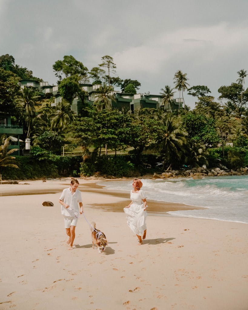 A couple walks their dog along a scenic beach in Phuket, Thailand, surrounded by lush greenery.