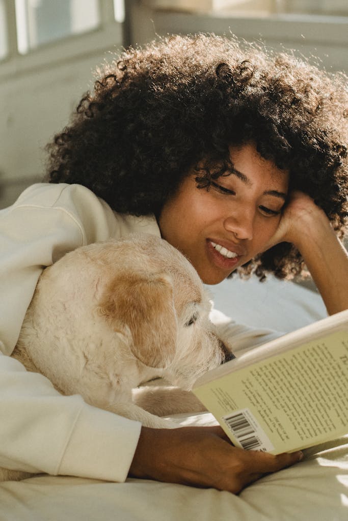 A joyful woman reads a book while relaxing indoors with her dog, embracing peaceful leisure time.