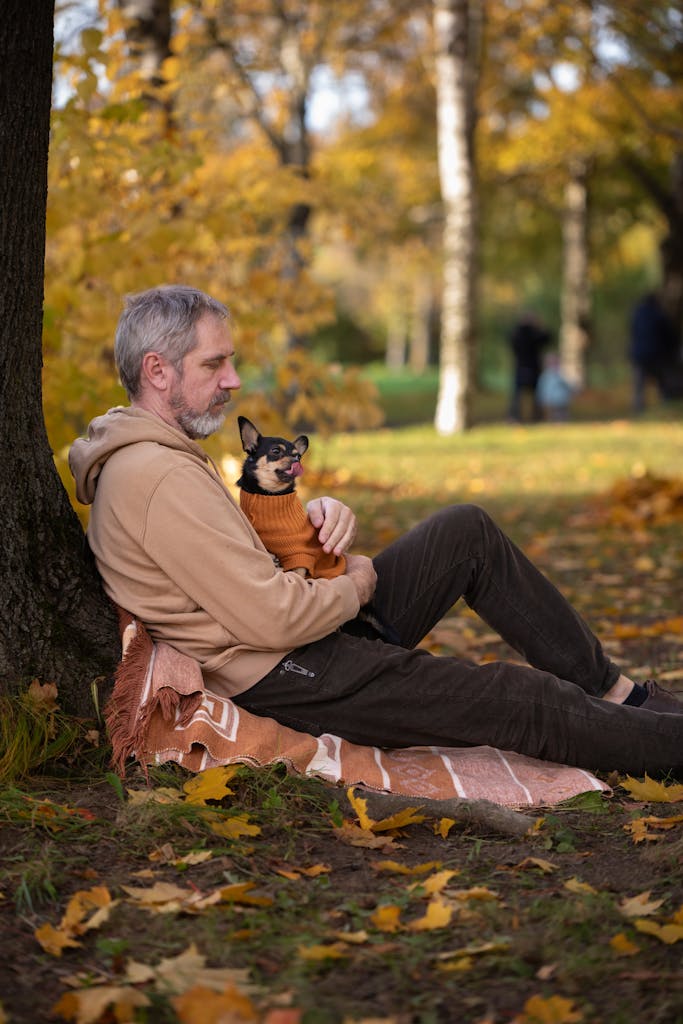 A man sitting by a tree holding his dog on a blanket during an autumn day in the park.