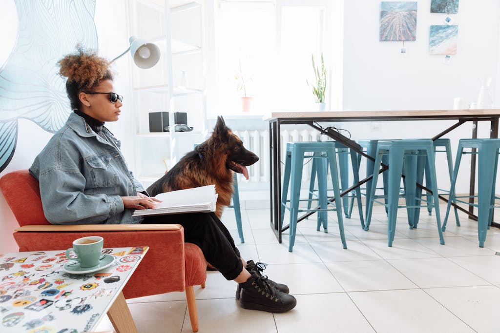 A visually impaired woman sits reading a braille book beside her guide dog indoors.