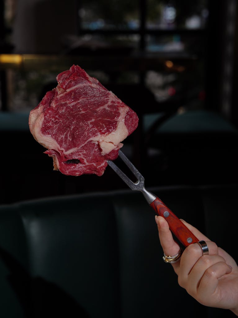 Close-up of a marbled ribeye steak on a fork, held by a hand, indoors in a restaurant setting.