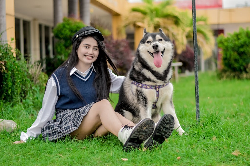 Young woman in a uniform sitting with a happy Siberian Husky on a vibrant green lawn.